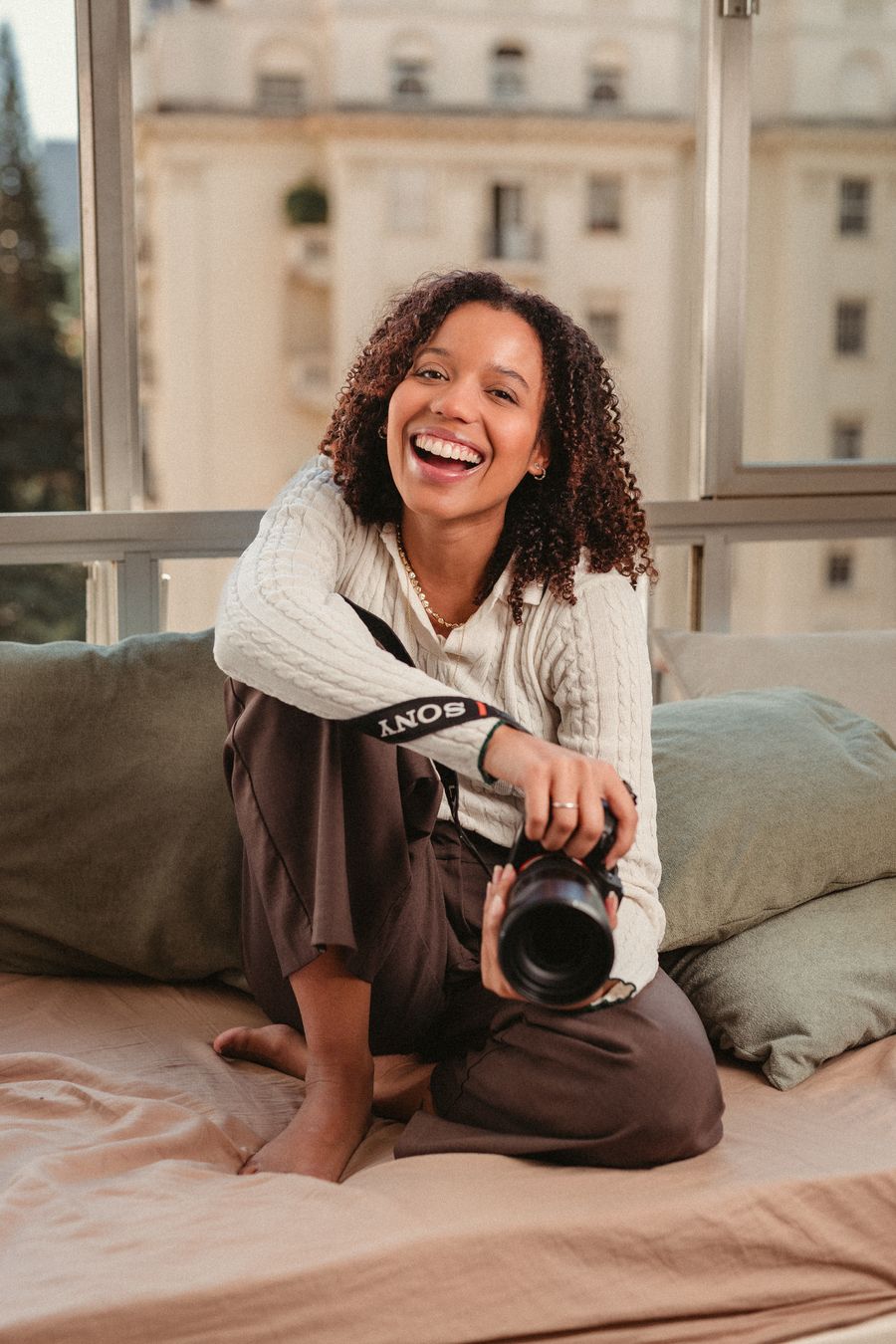 Isadora Hadassa sorrindo, sentada junto à janela com a câmera na mão